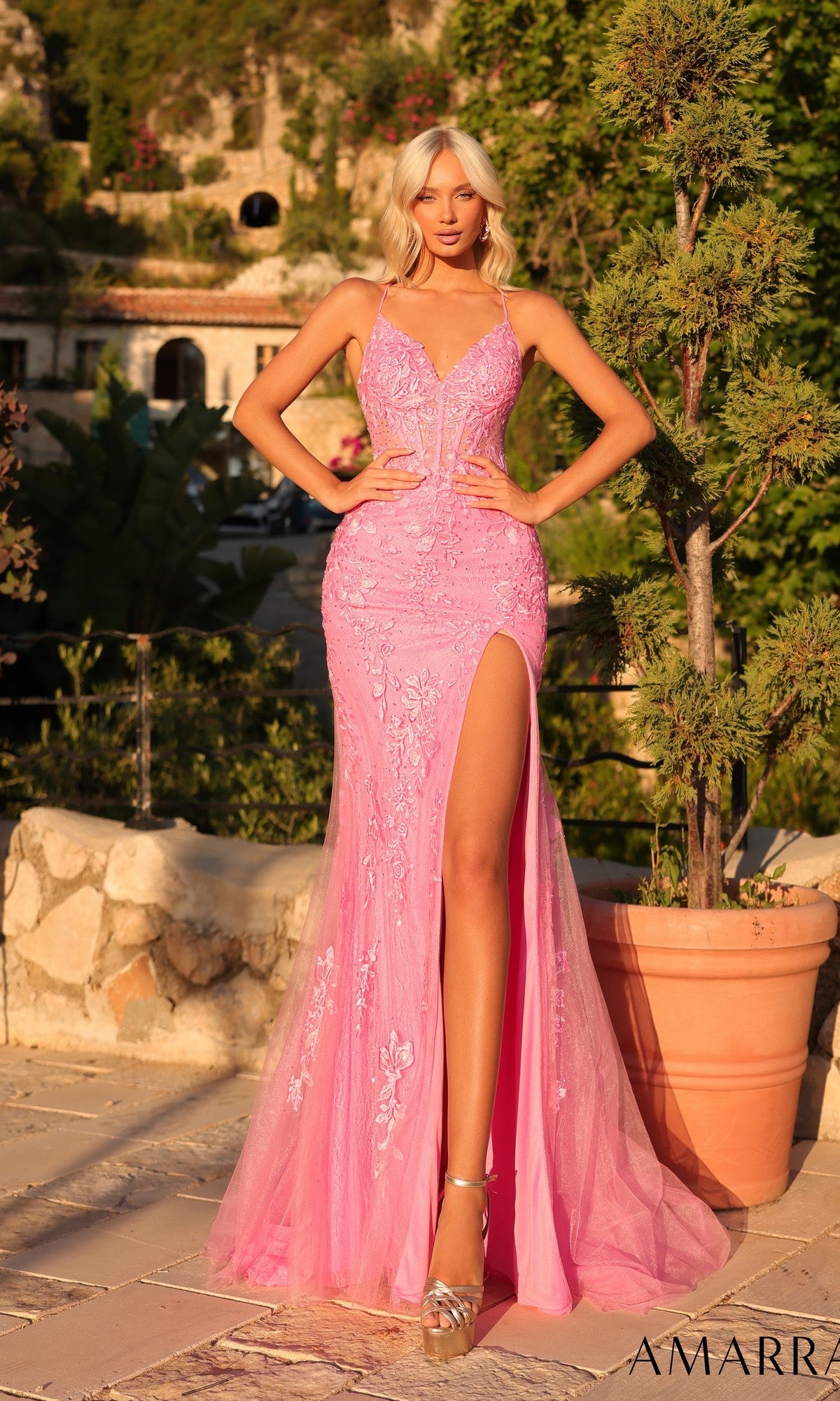 A woman wearing the Embroidered-Lace Long Prom Dress: Amarra 89022, featuring pink embroidery, strappy mermaid silhouette, and a thigh-high slit, stands outdoors on a stone patio by a potted plant.
